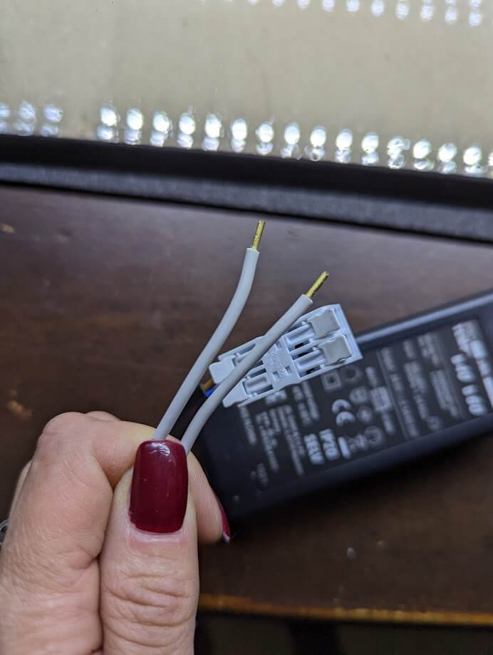 Eine Hand mit roten Fingernägeln hält zwei graue, abisolierte Kabel und einen grauen Stecker neben einem schwarzen Netzteil