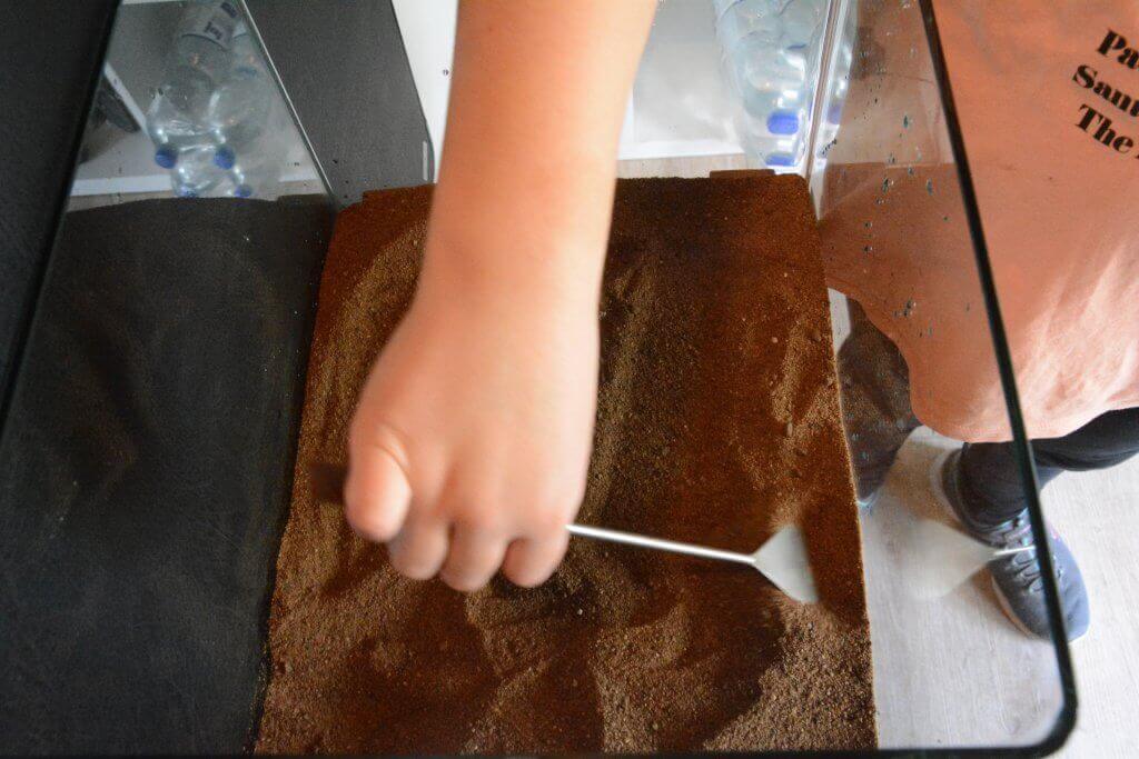 Eine Hand glättet mit einem Metallwerkzeug braunes Substrat im leeren Aquarium; im Hintergrund stehen Wasserflaschen